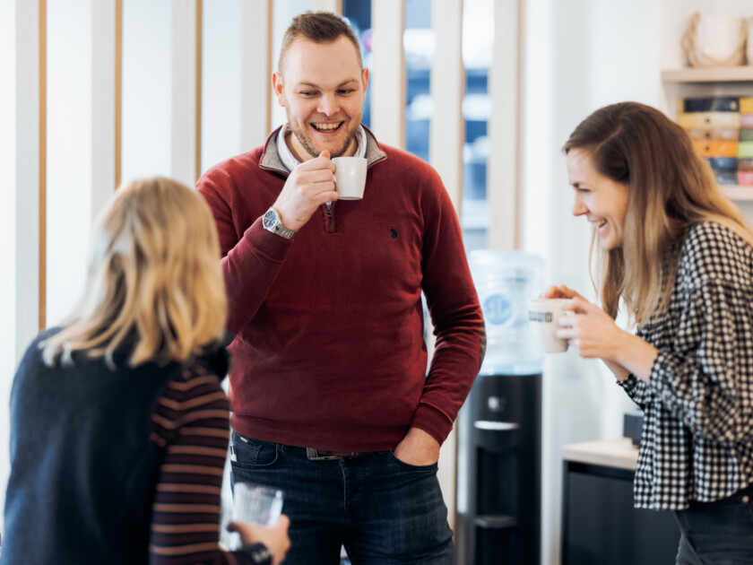 Three colleagues chatting and smiling in modern office kitchen during coffee break