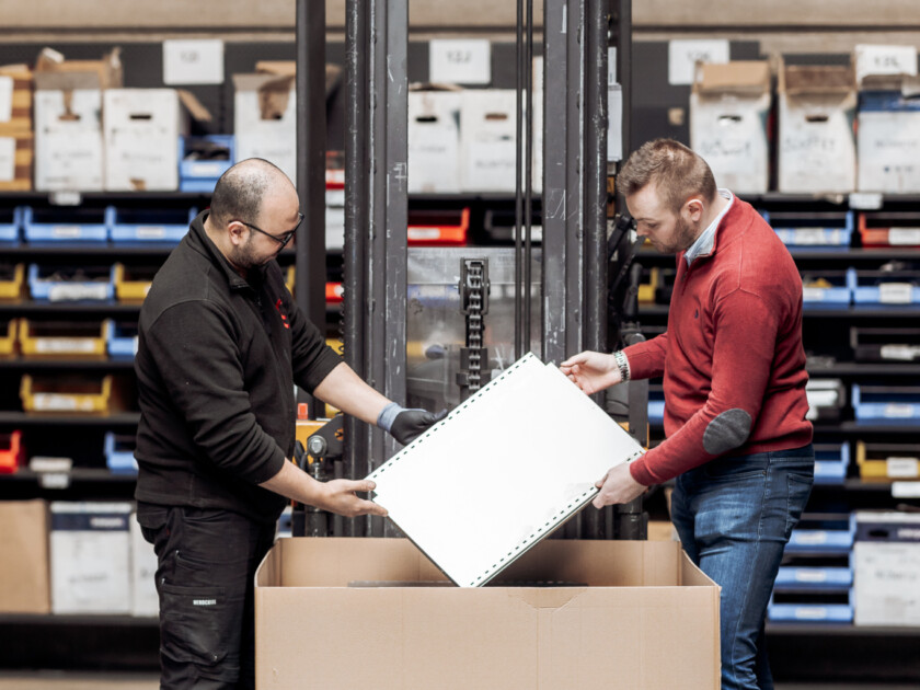 Two warehouse workers inspecting white document or product specification sheet over large Gondella branded cardboard box with forklift in background
