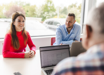 Two professionals in office setting discussing retail solutions with laptops, overlooking parking lot with vehicles