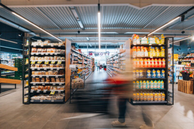 Delhaize supermarket with Pitch shelving displaying packaged goods and beverages in central aisle with metal-framed display units