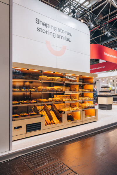 Gondola bakery display with warm lighting, orange interior, and 'Shaping stores, storing smiles' tagline at Euroshop 2023