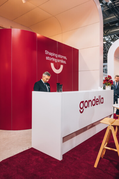 Gondella exhibition booth at EuroShop 2023 with red curved wall, white reception counter, and smiley logo tagline 'Shaping stores, storing smiles'