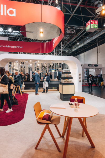Gondella retail booth at Euroshop 2023 with curved red branding, white service counter, wooden table with orange chair, and red carpet flooring.