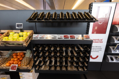 Wine display with black bottle rack shelving, yellow apples and oranges in wooden crates, and digital price promoter in modern supermarket