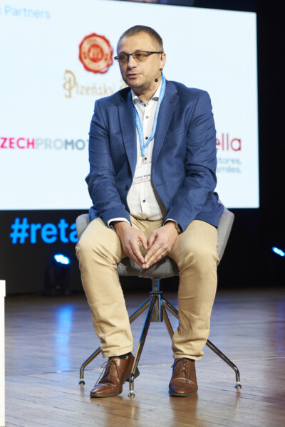 Speaker presenting at retail industry conference on stage with event branding visible
