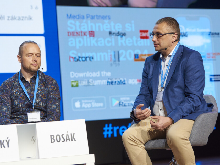 Two speakers at retail conference panel discussion with event backdrop displaying media partners and summit branding
