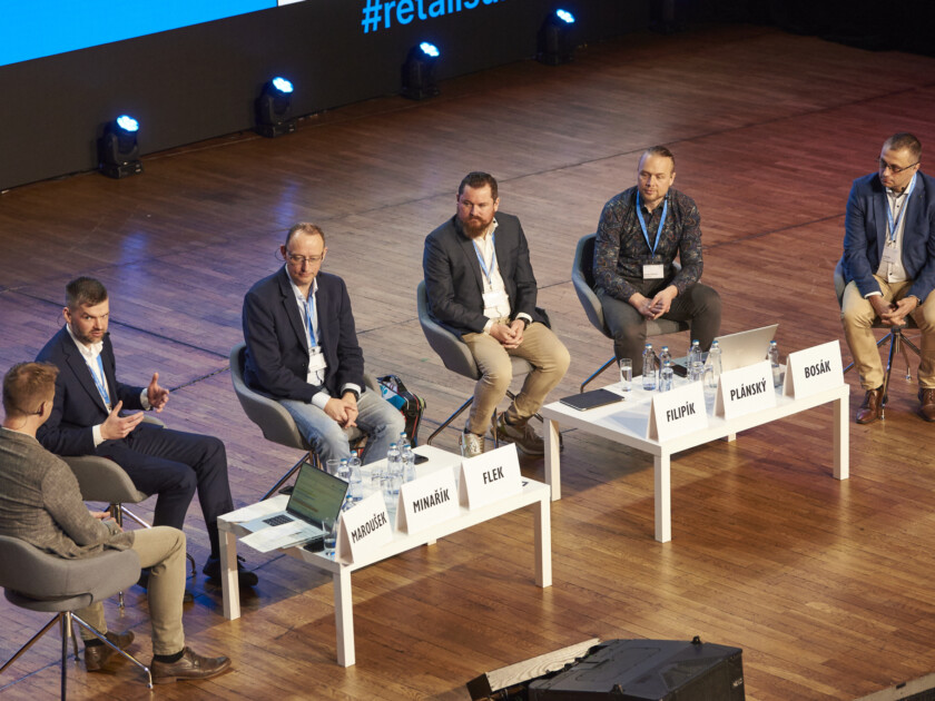 Panel discussion at retail industry conference with five speakers seated at white table on wooden stage