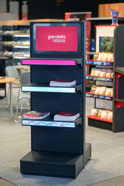 Gondella digital display kiosk with shelving, magenta branding band, and product literature in retail environment