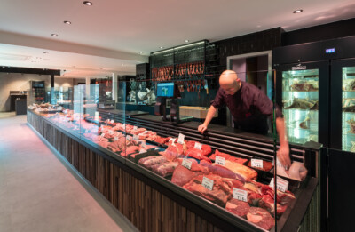 Butcher shop display with refrigerated meat counter, glass-fronted cases showing cuts of beef and poultry, staff member arranging products, neon signage overhead