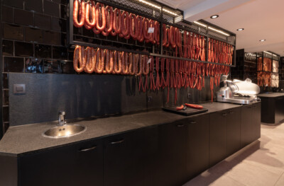 Specialty food retail display with cured meat products hanging on wall-mounted mesh shelving, dark tile backsplash, and stainless steel counter with sink