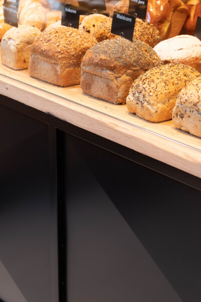 Artisan bread loaves with seed toppings displayed on wooden shelf with product name labels in bakery retail setting