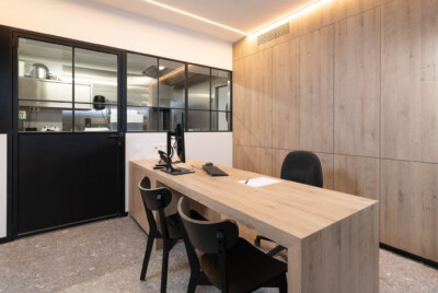 Modern office workspace with light wood wall paneling, black-framed glass partition, wooden desk with computer monitor and keyboard, black office chairs
