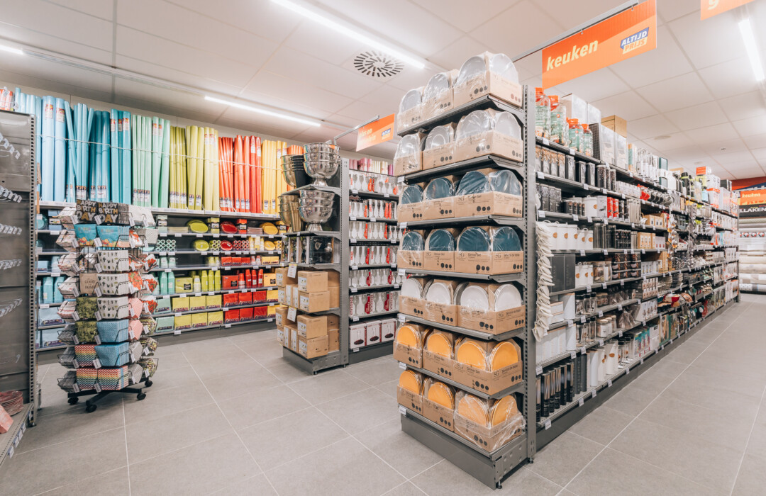 Discount supermarket interior with Pitch shelving displaying kitchenware, colorful curtains, baskets on rotating display, and packaged goods on wall-mounted gondolas