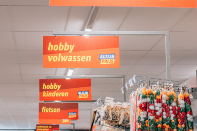 Hobby section signage with Altijd Prijs labels for volwassen, kinderen, and fietsen categories in Yess Waregem store
