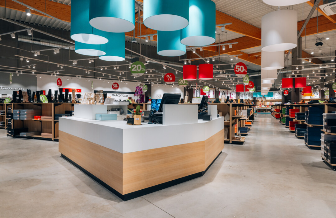 Modern retail showroom with central checkout counter, Pitch shelving displays, suspended cylindrical lighting, and promotional signage in supermarket setting