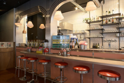 Modern horeca bar counter with red vinyl stools, white subway tile backsplash, pendant lighting, and stocked shelving