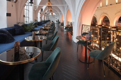 Modern horeca lounge with arched ceilings, blue seating, round tables with brass bases, and decorative railings in contemporary restaurant setting