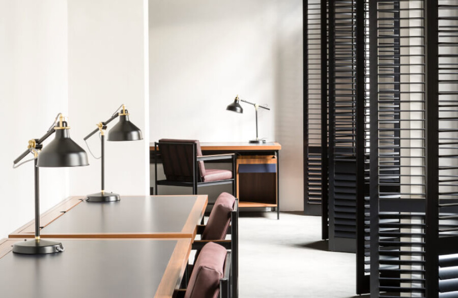 Modern office showroom with wooden desks, adjustable task lamps, pink upholstered chairs, and black louvered room dividers