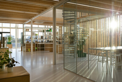 Modern office interior with glass partition walls, wooden shelving units, and natural lighting from floor-to-ceiling windows