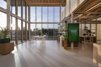 Modern retail showroom with wooden shelving systems, display counter with green screen, and floor-to-ceiling windows overlooking landscape