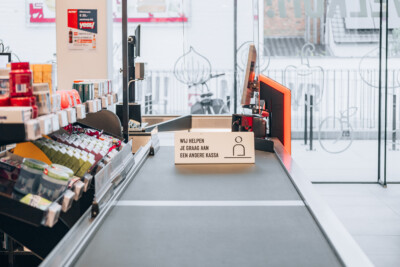 Delhaize checkout counter with red signage directing customers to another register, displaying product samples and retail fixtures