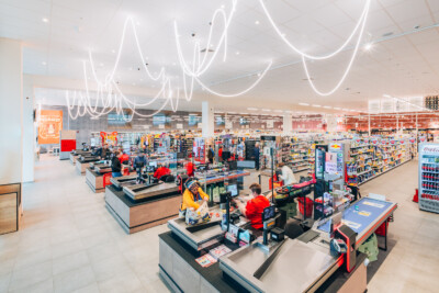 Modern supermarket checkout area with Pitch shelving displaying groceries, beverages, and products across multiple aisles with white neon lighting