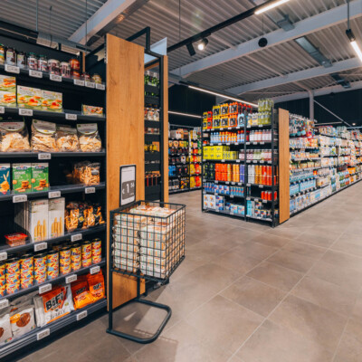 Pitch 25 shelving with orange and yellow packaged goods in supermarket aisle, featuring wooden-framed end cap display and metal basket stand