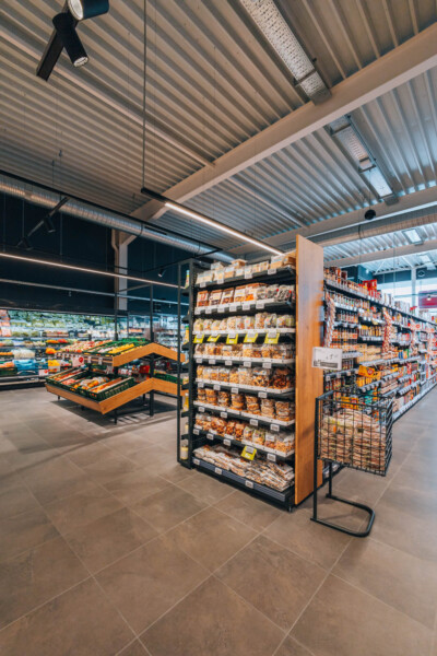 Delhaize supermarket interior with Pitch shelving displaying packaged goods, produce display, and modern retail layout under corrugated ceiling