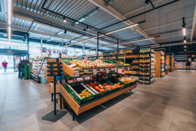 Delhaize supermarket produce section with Pitch 25 shelving displaying fresh fruits and vegetables, packaged goods shelving visible in background