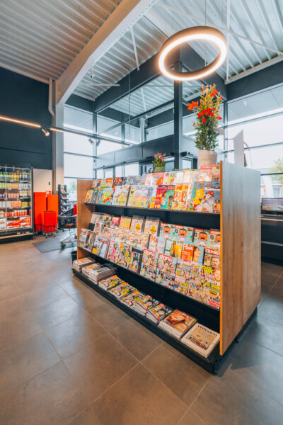 Pitch shelving display with magazines and publications in supermarket checkout area, topped with potted flowers and neon ring light