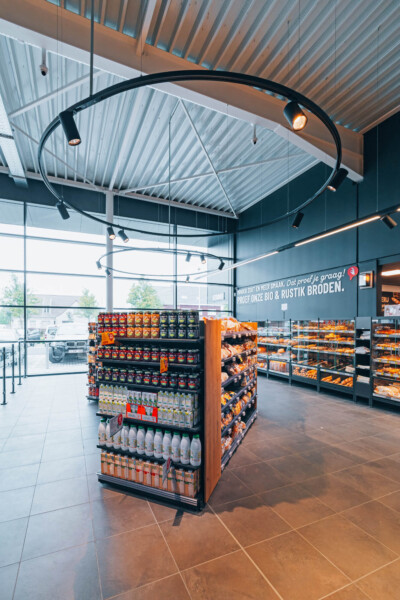 Delhaize supermarket interior with central Pitch 41 shelving display unit, orange and black product packaging, modern ceiling with skylight, and wall-mounted shelving in background