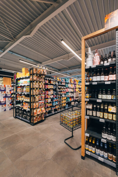 Delhaize supermarket interior with Pitch shelving displaying groceries, beverages, and wine in organized retail layout under industrial ceiling
