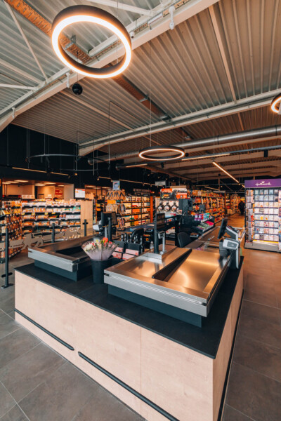 Delhaize supermarket checkout area with orange neon ring lights, wooden counter, shelving displays, and corrugated metal ceiling
