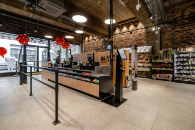 Delhaize supermarket checkout area with wooden Pitch shelving, POS terminals, red decorations, and refrigerated displays