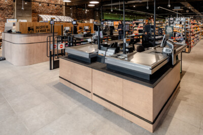 Proxy Delhaize supermarket checkout area with wooden checkout counters, shelving displays, and brick accent wall