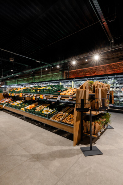 Produce display with green Pitch shelving units holding fresh vegetables and oranges in a supermarket, with brown paper bag dispensers and refrigerated cases in background