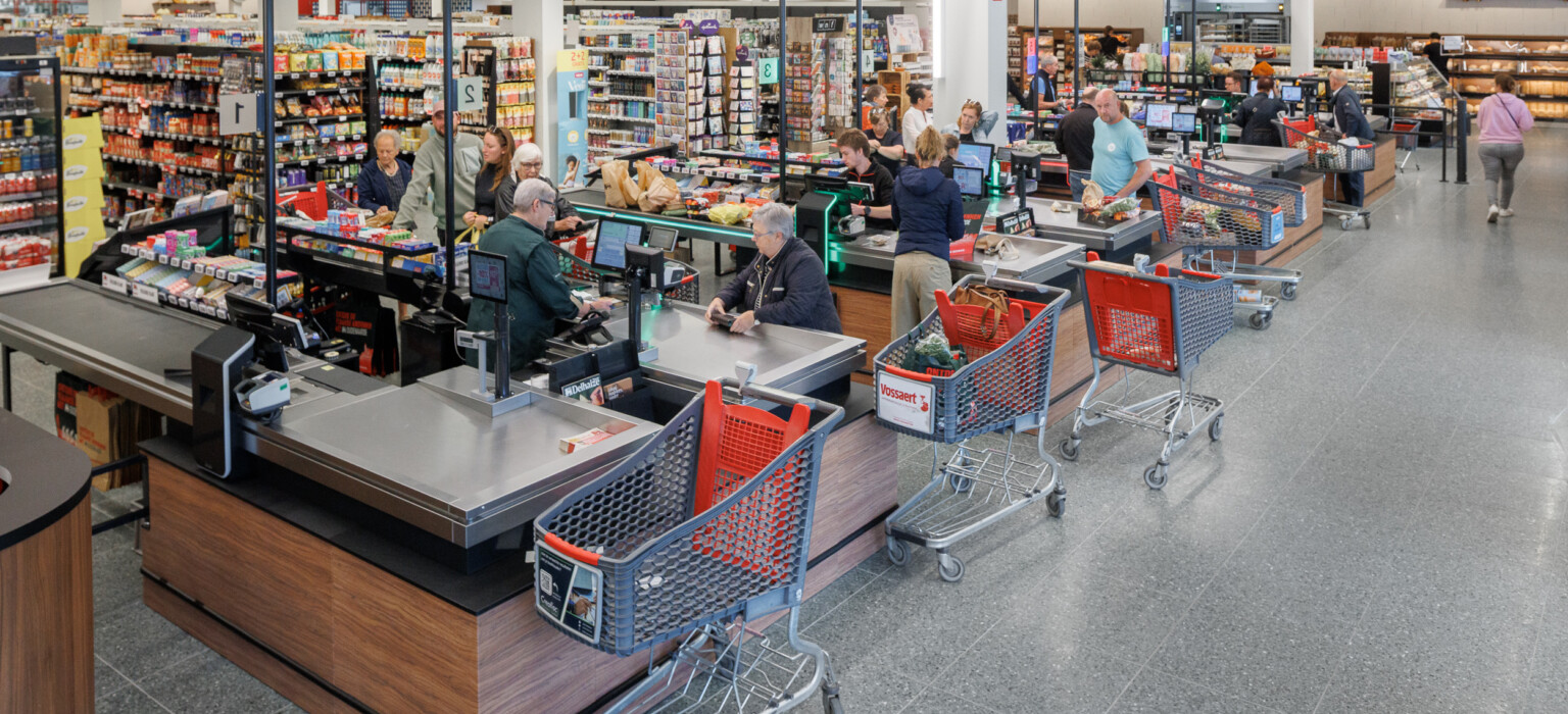 Delhaize supermarket checkout area with multiple Gondella counters, red shopping carts, and stocked shelving in background
