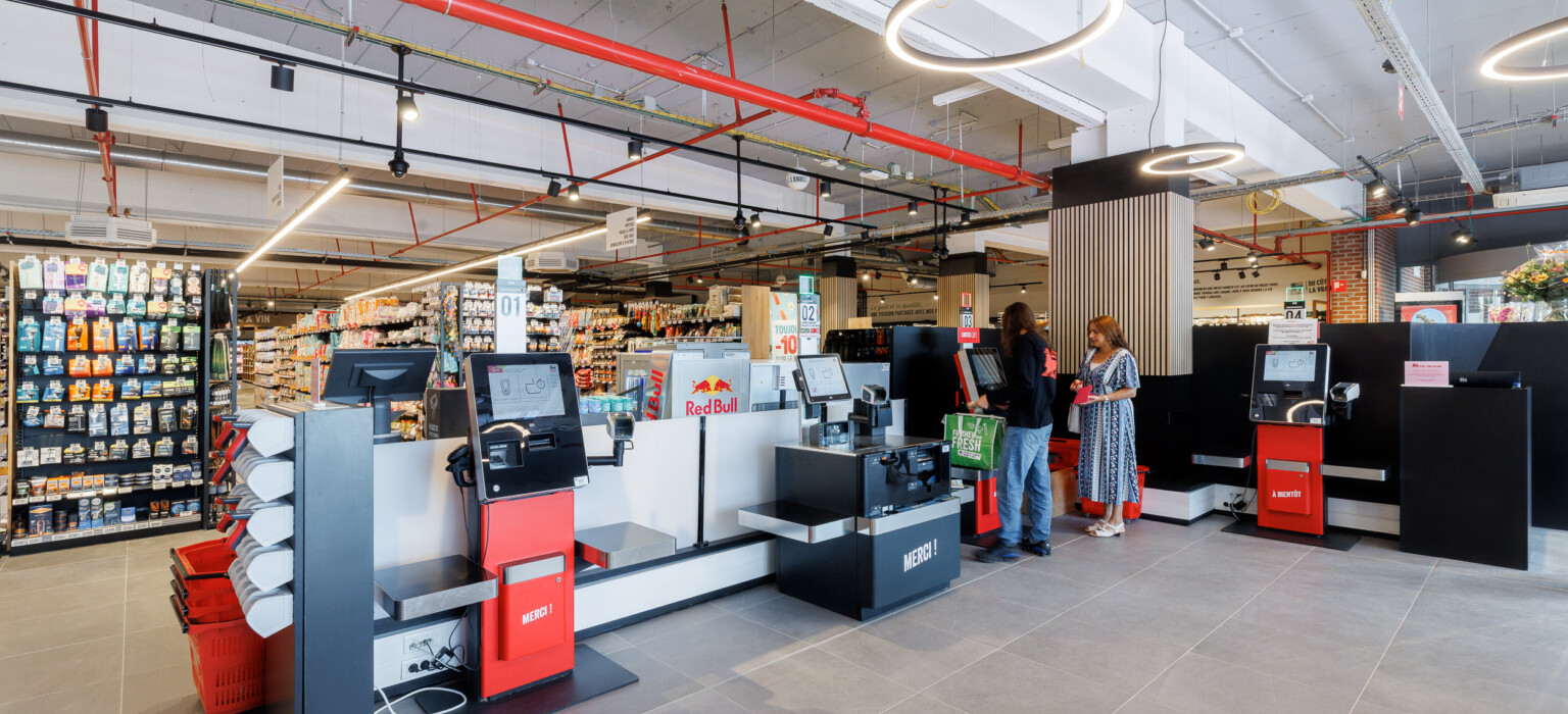 Delhaize supermarket checkout area with red and black Gondella counters, shelving displays, and modern ceiling lighting