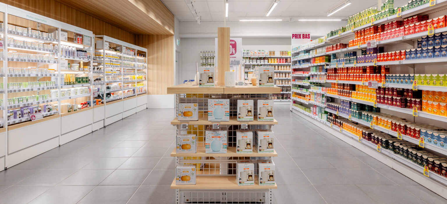 Delhaize supermarket aisle with Pitch shelving displaying health & beauty and grocery products, central display stand with wooden shelves