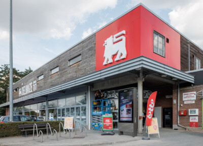 Delhaize supermarket storefront with red signage, Pitch 41 shelving visible through windows, and outdoor seating area
