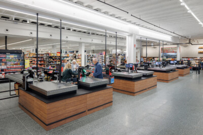 Delhaize supermarket checkout area with wooden counter fixtures, glass partition screens, and stocked shelving in background