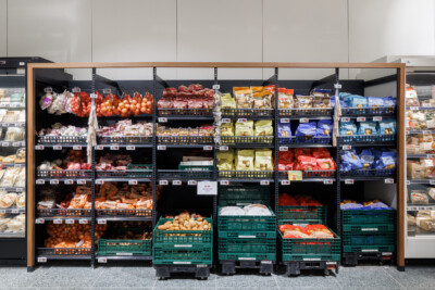 Delhaize supermarket produce and meat display with Pitch 25 shelving, fresh vegetables in mesh bags, packaged meats, and organized retail layout