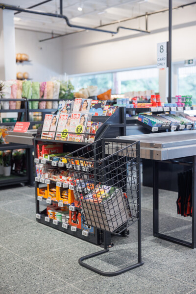 Delhaize supermarket checkout area with black metal cart display stand and magazines, colorful product shelving visible in background