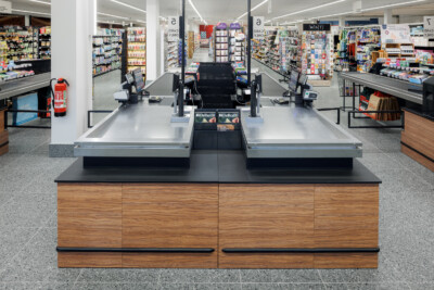 Delhaize checkout counter with dual POS stations, black countertop and wood-grain base, supermarket interior with stocked shelving visible