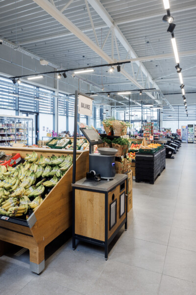 Delhaize supermarket produce section with Gondella wooden shelving displaying bananas, oranges and fresh fruits under industrial ceiling