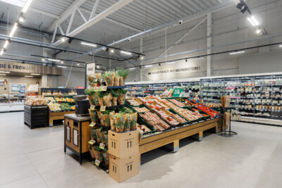 Delhaize supermarket produce section with tiered wooden display tables, fresh vegetables and fruit, refrigerated cases, and Pitch shelving in background