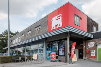 Delhaize supermarket storefront with red signage, Pitch 41 shelving visible through windows, and outdoor seating area
