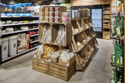 Wooden crate display with packaged specialty food products in specialty food retail store, featuring angled shelving and refrigerated cases